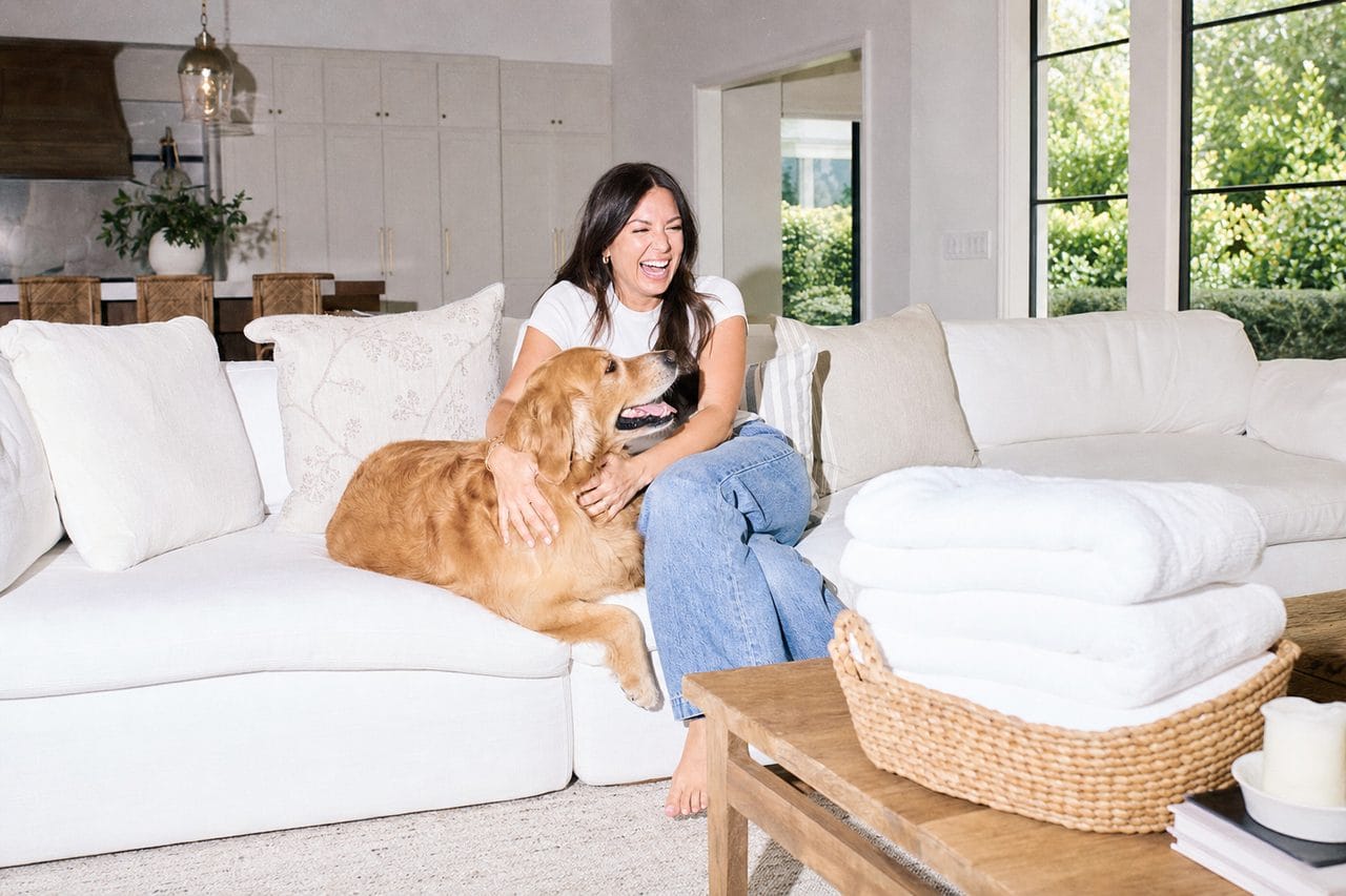 A bright clean living room, a woman laughing on the sofa with her dog